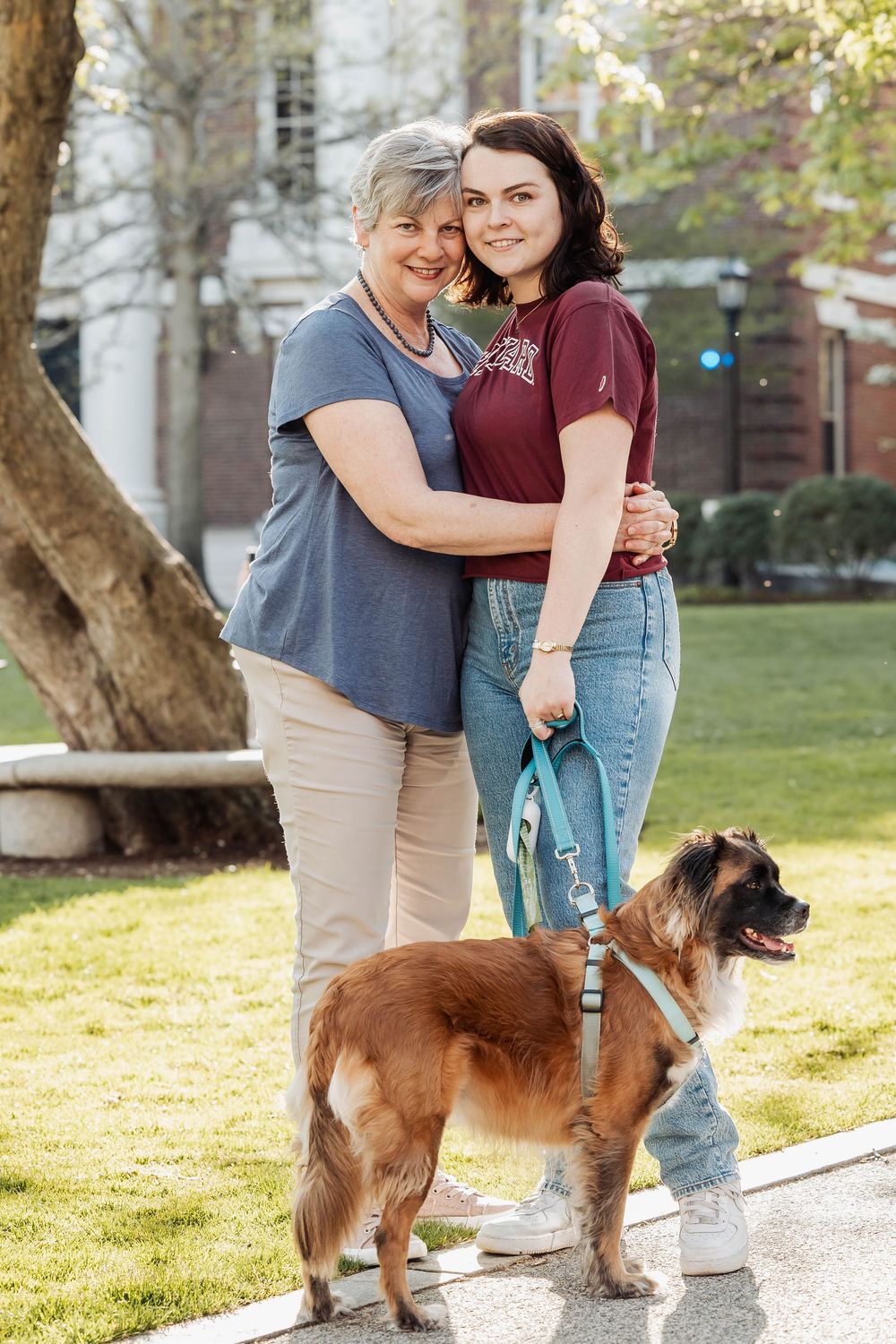 A dog on a leash stands next to two people embracing on a sunny sidewalk beside a grassy lawn.