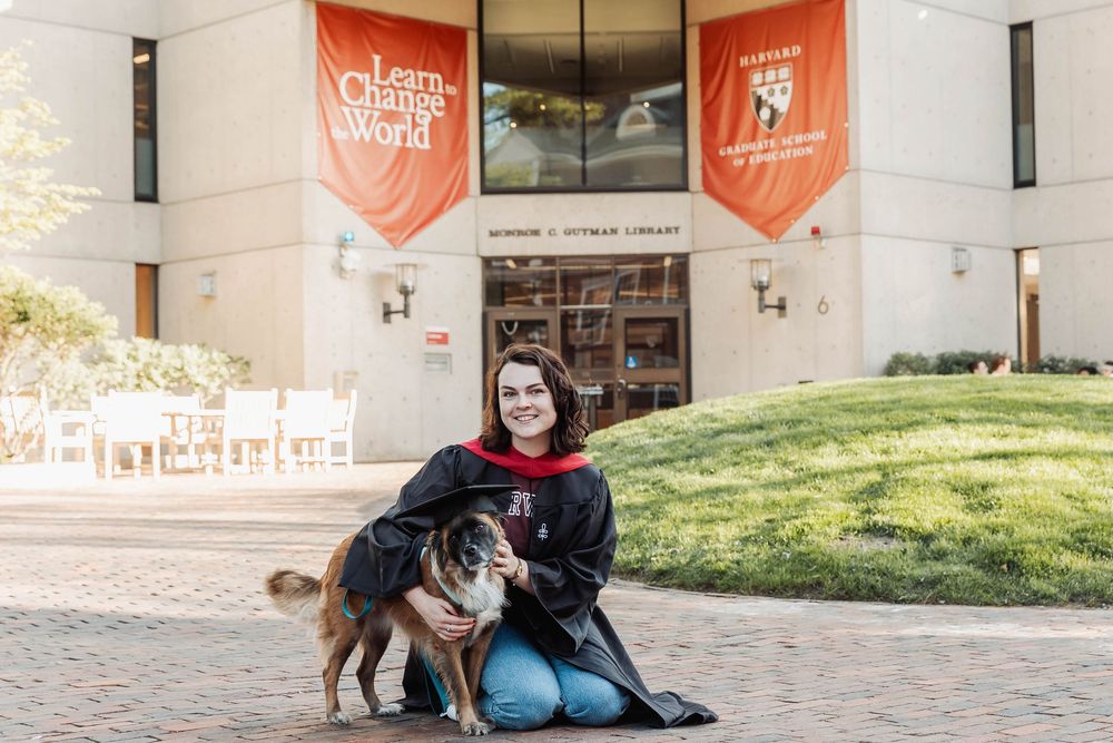 A person sits with two dogs outside a building with orange banners at the Bowling Green State University campus entrance.