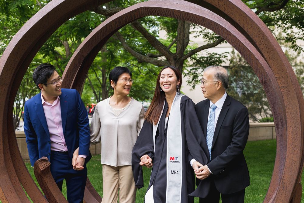 A graduate poses with family members under a circular metal arch structure in a green outdoor setting.