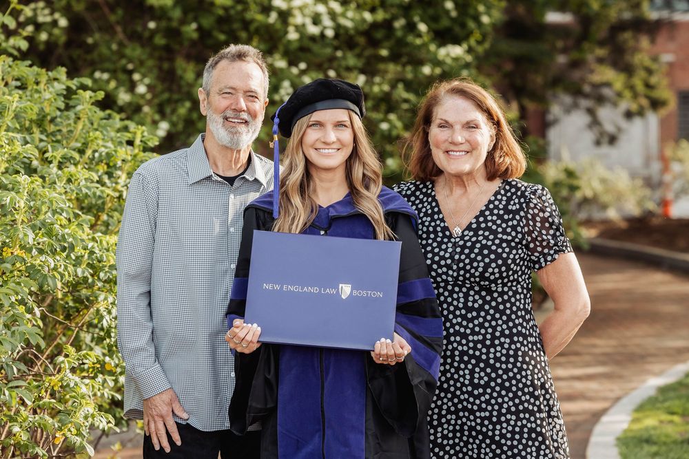 A graduate in blue cap and gown holds a diploma while posing with two supporters on a tree-lined pathway.
