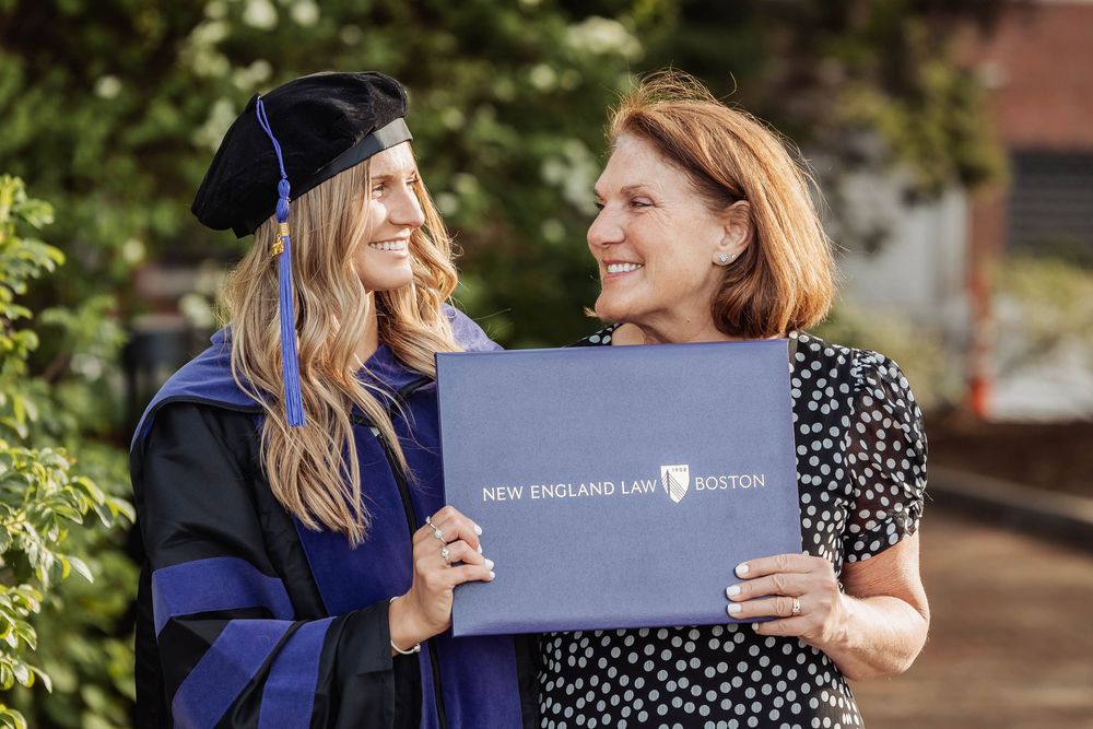 A graduate in academic regalia holds their graduation diploma folder while sharing a joyful moment outdoors.