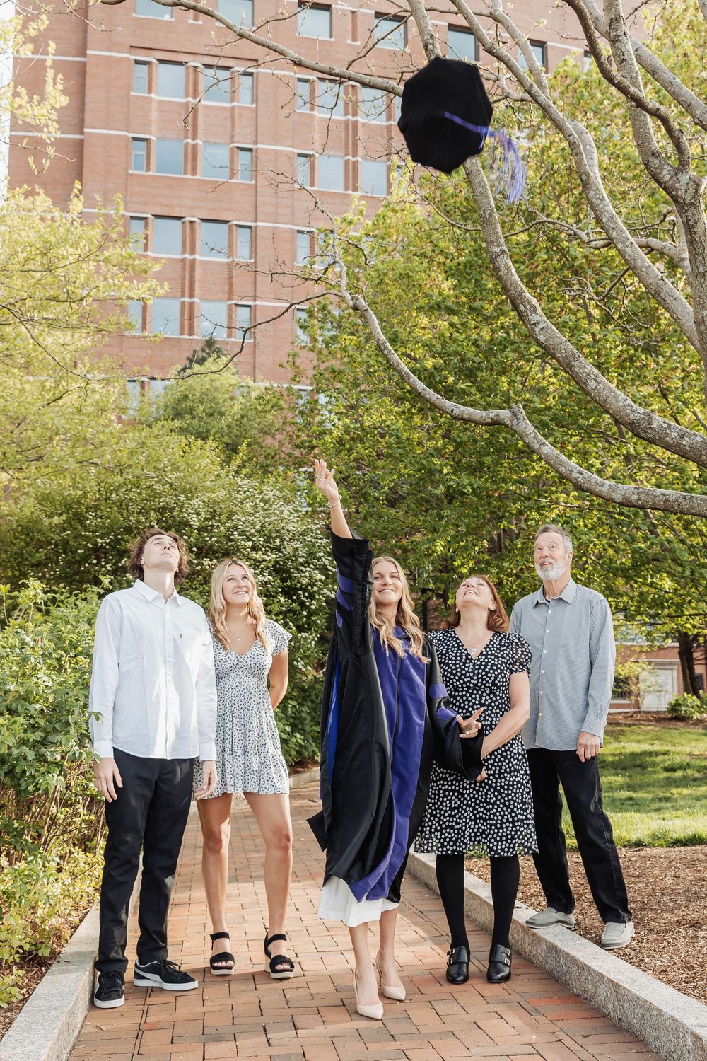 Graduate celebrates by tossing cap in air while standing with family on brick pathway near brick building and trees.