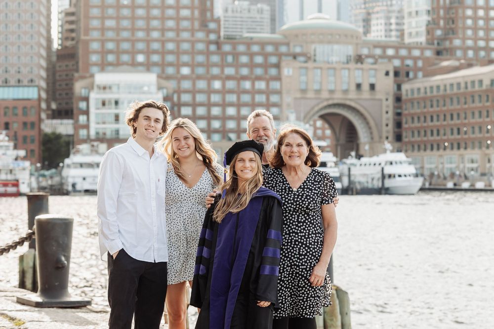 A group stands together smiling by a waterfront with city buildings and boats in the harbor behind them.