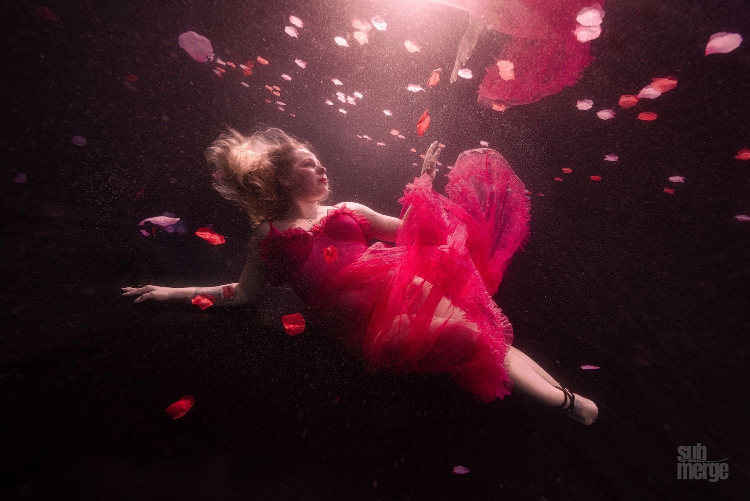 Figure in flowing red dress dances gracefully underwater surrounded by floating rose petals in dark dramatic lighting.