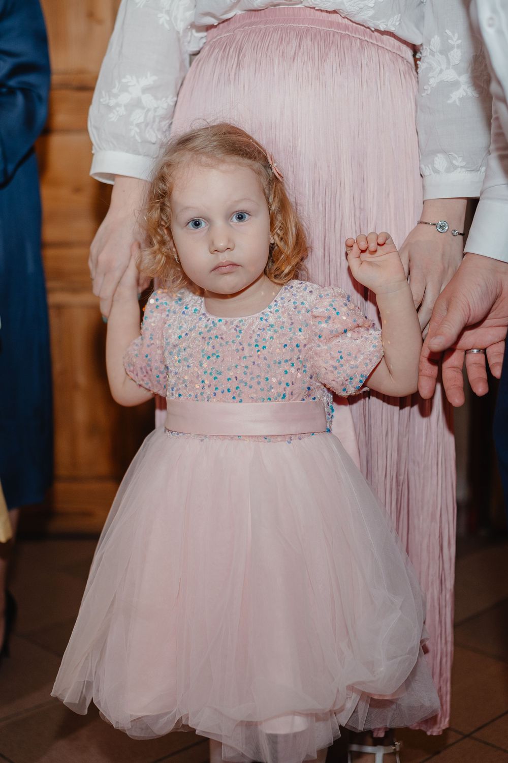 A child wearing a pale pink tulle party dress with floral details stands holding someone's hand at an indoor event.
