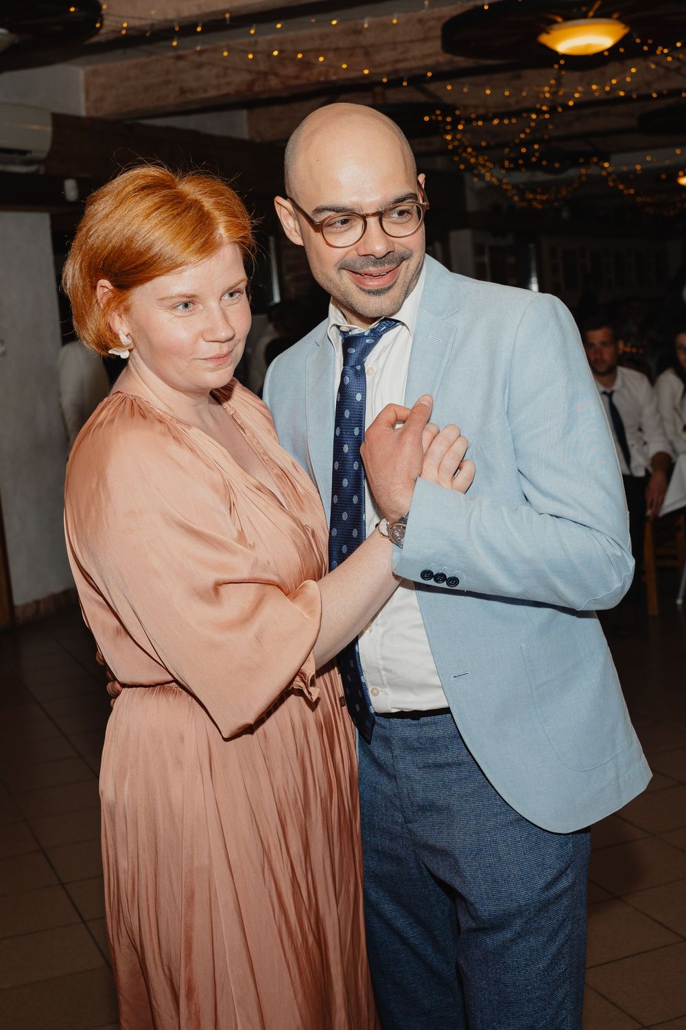 A couple poses together at an event, wearing a peach dress and light blue blazer with navy tie beneath twinkling lights.