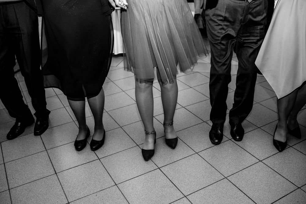 Black and white photograph showing three pairs of legs wearing formal shoes standing on tiled floor at gathering.
