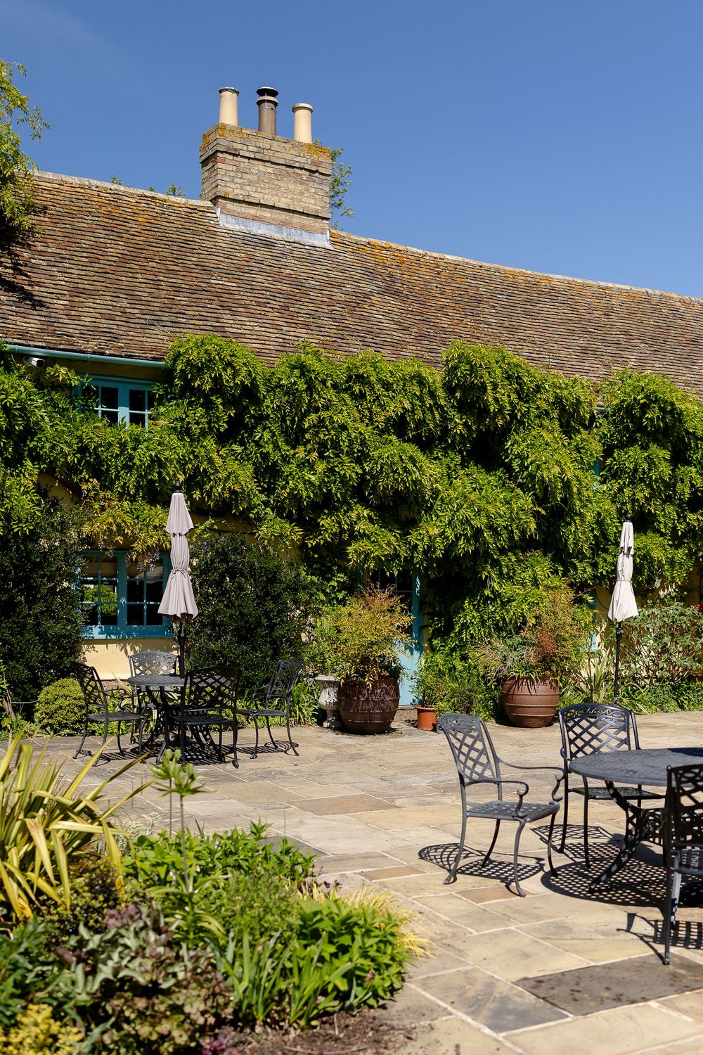 Outdoor patio seating area with wrought iron furniture against a historic cottage covered in climbing vines.