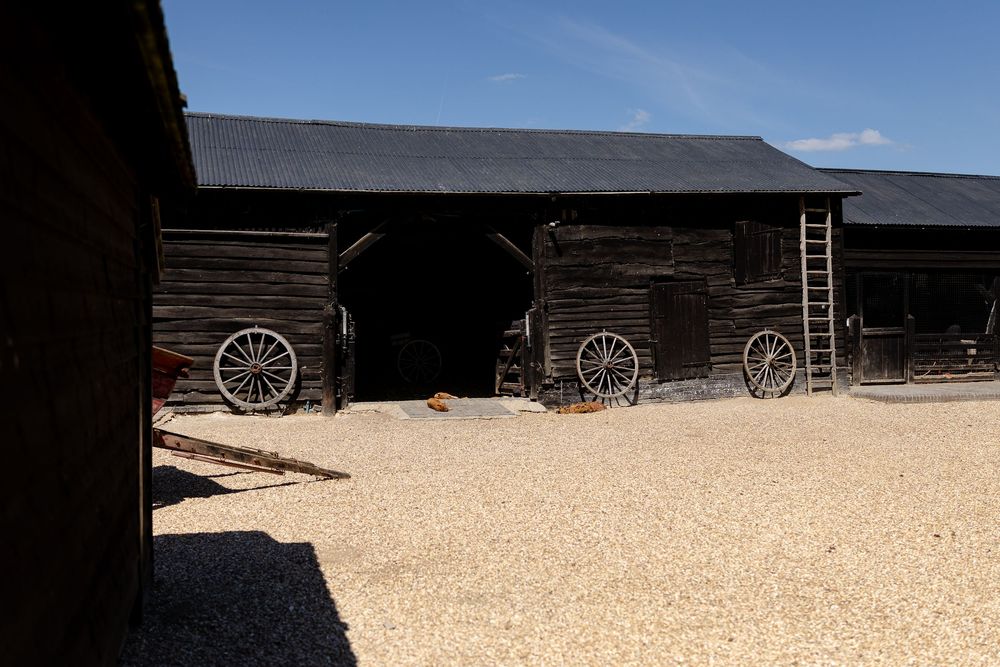 Rustic wooden barns with wagon wheels leaning against dark weathered walls on a gravel courtyard under blue sky.