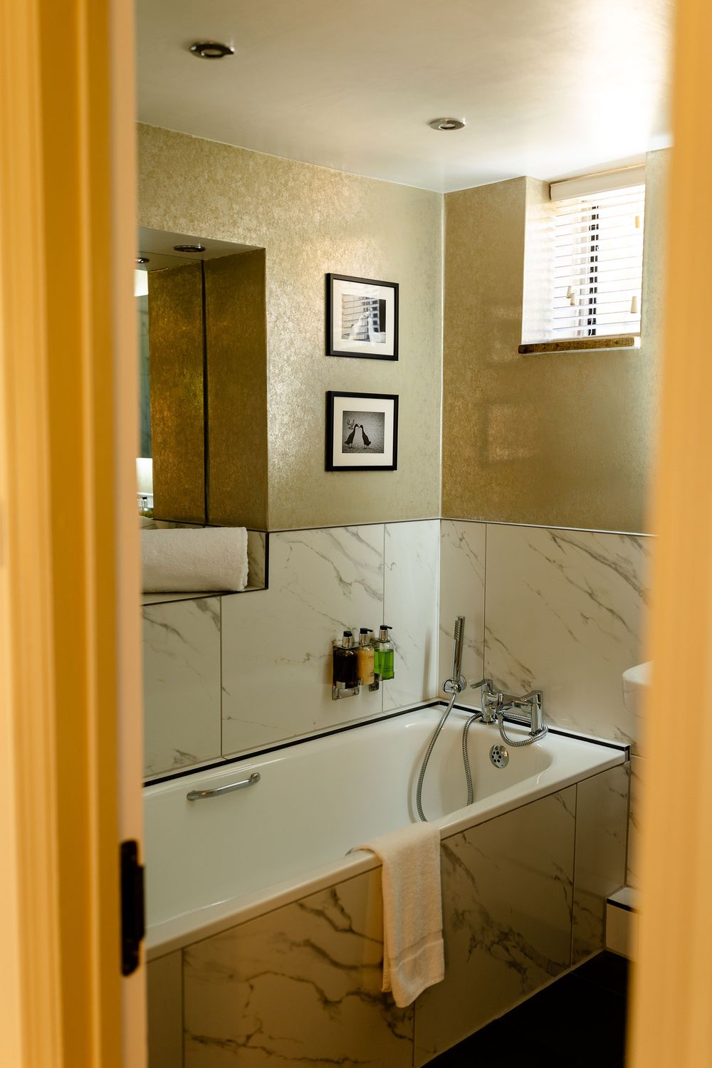 Luxurious marble bathroom with soaking tub, gold walls, framed artwork and hotel amenities displayed on tub ledge.
