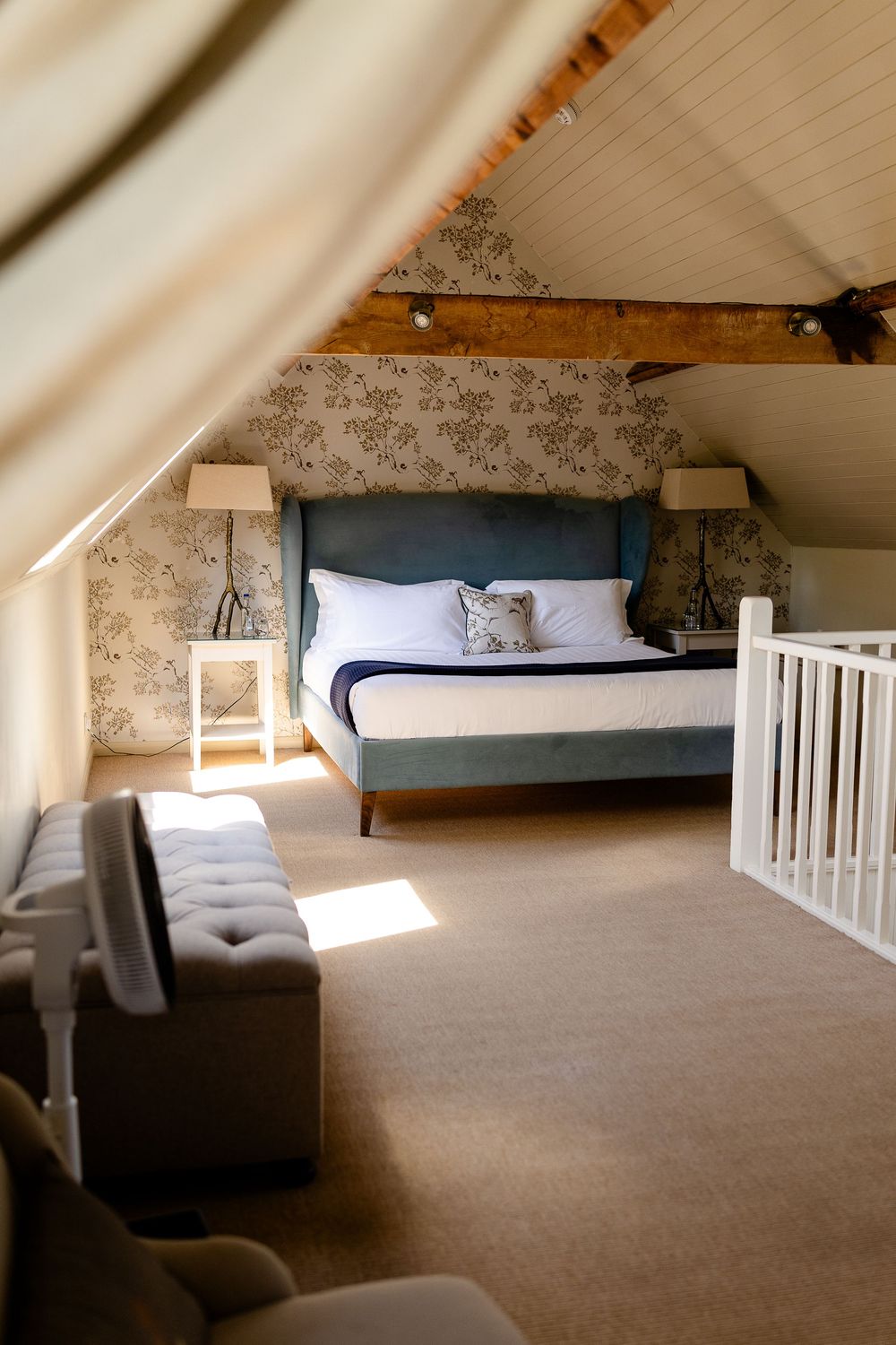 A cozy attic bedroom with a blue bed, floral wallpaper, exposed wooden beams, and sunlight streaming through a window.