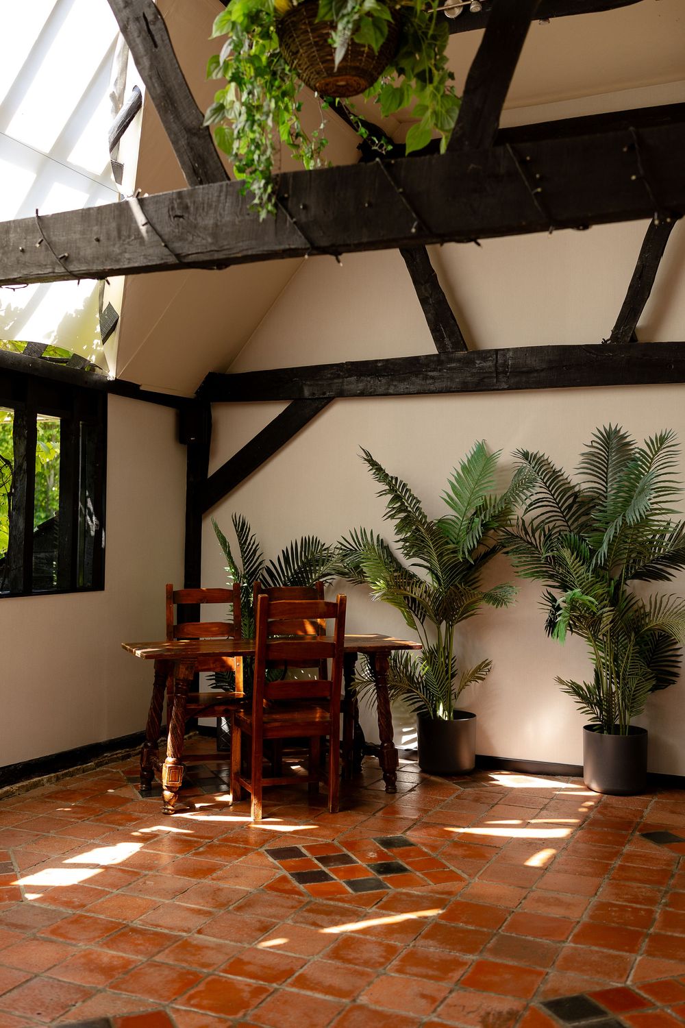Rustic interior space with exposed wooden beams, terracotta flooring, and potted palm plants near wooden furniture.