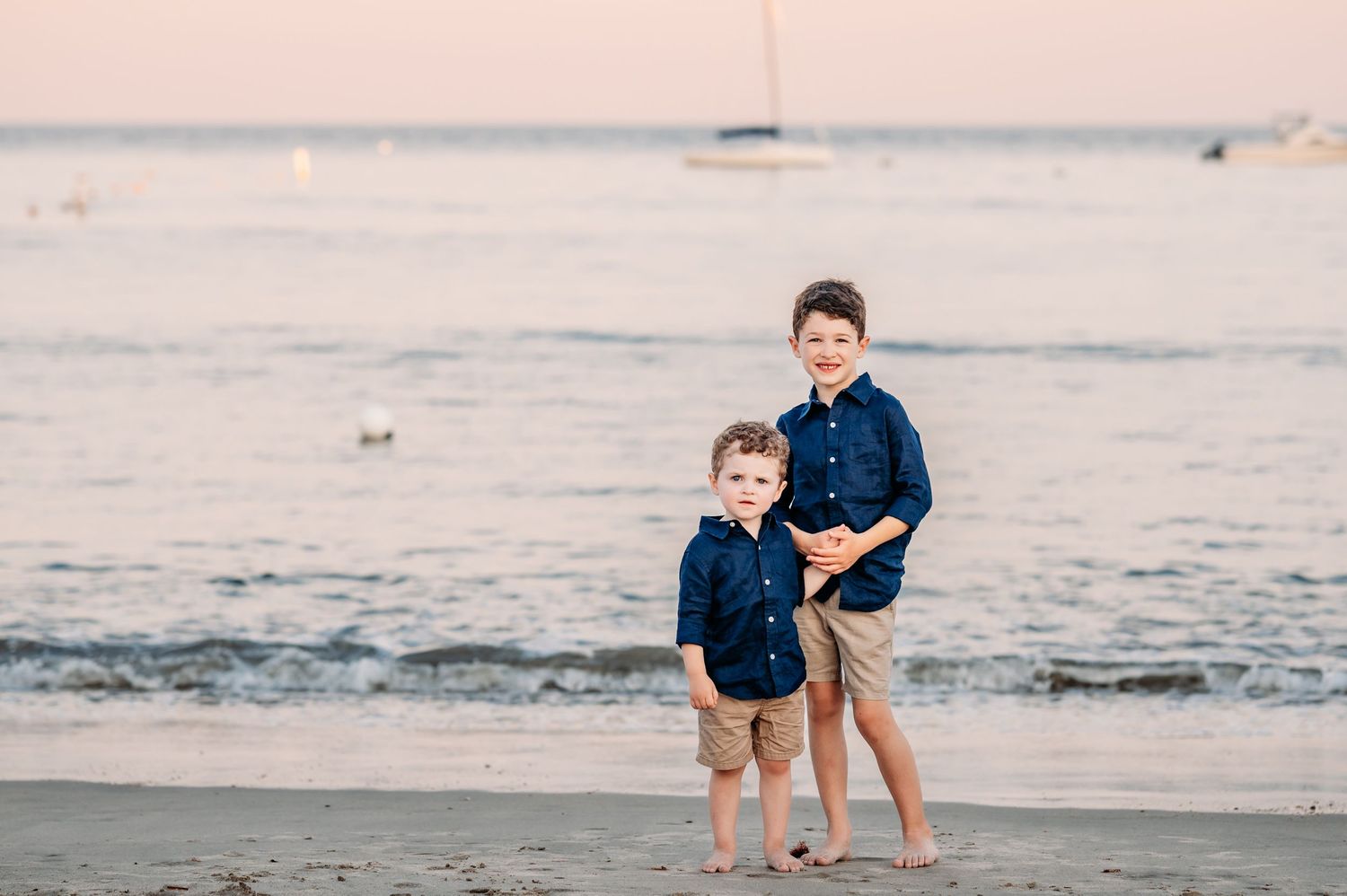 Two children in matching navy shirts standing together on a beach at sunset with ocean waves in the background.