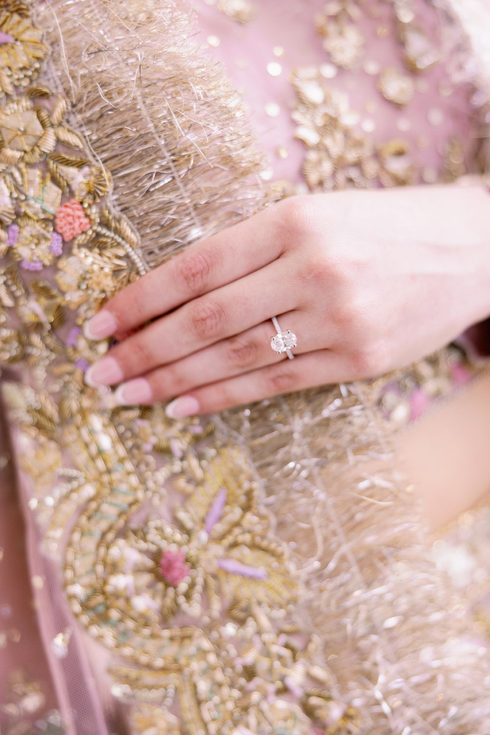 Close-up of a delicate engagement ring on hand against embellished pink and gold fabric.
