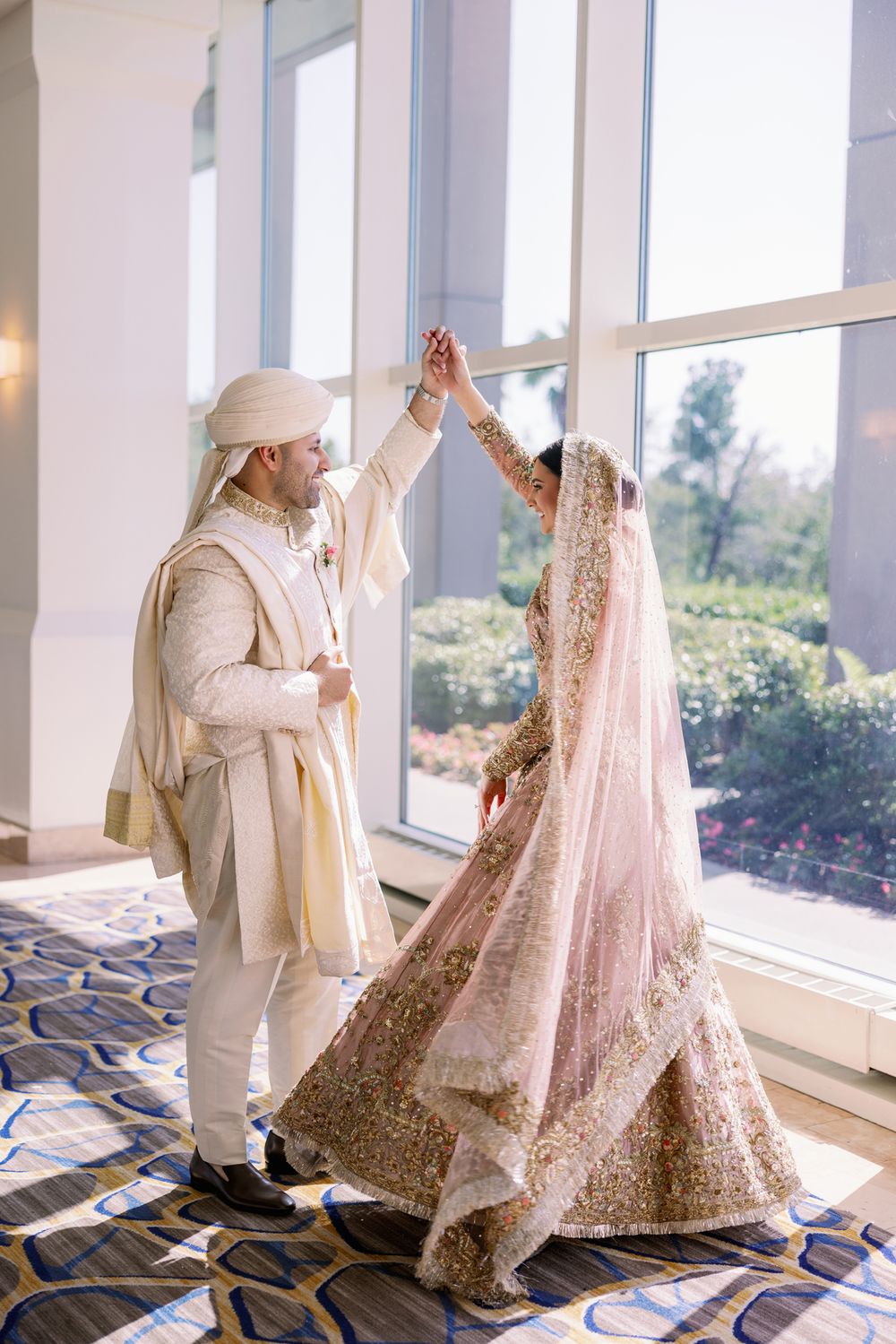 Couple in traditional South Asian wedding attire dancing together in front of large windows.