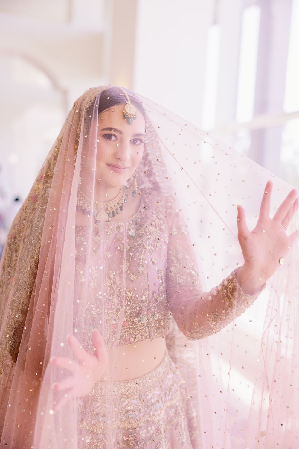 Bride in pink embellished lehenga poses behind sheer veil with sparkling details.