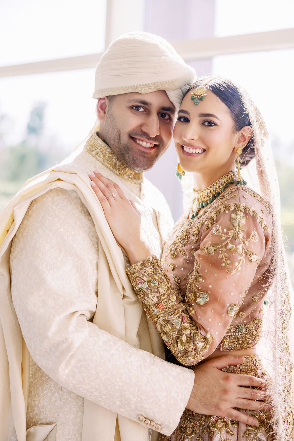 Newlyweds pose in elegant cream and pink traditional South Asian wedding outfits.