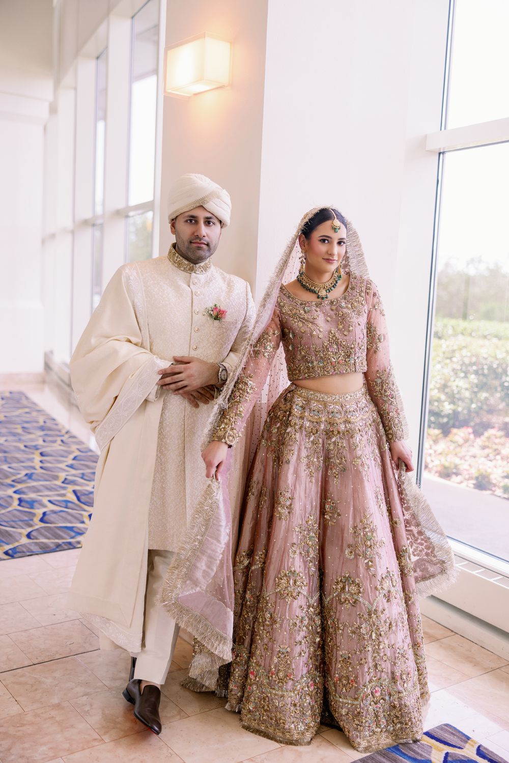 Couple in cream and pink South Asian wedding attire pose by bright windows.