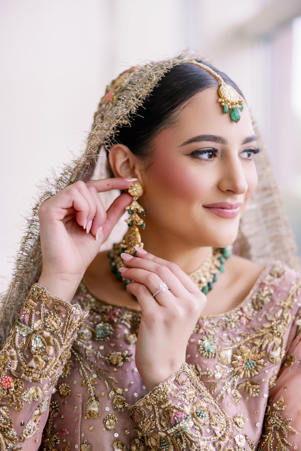 Bride in embellished pink lehenga adjusts her jewelry while wearing traditional bridal accessories.