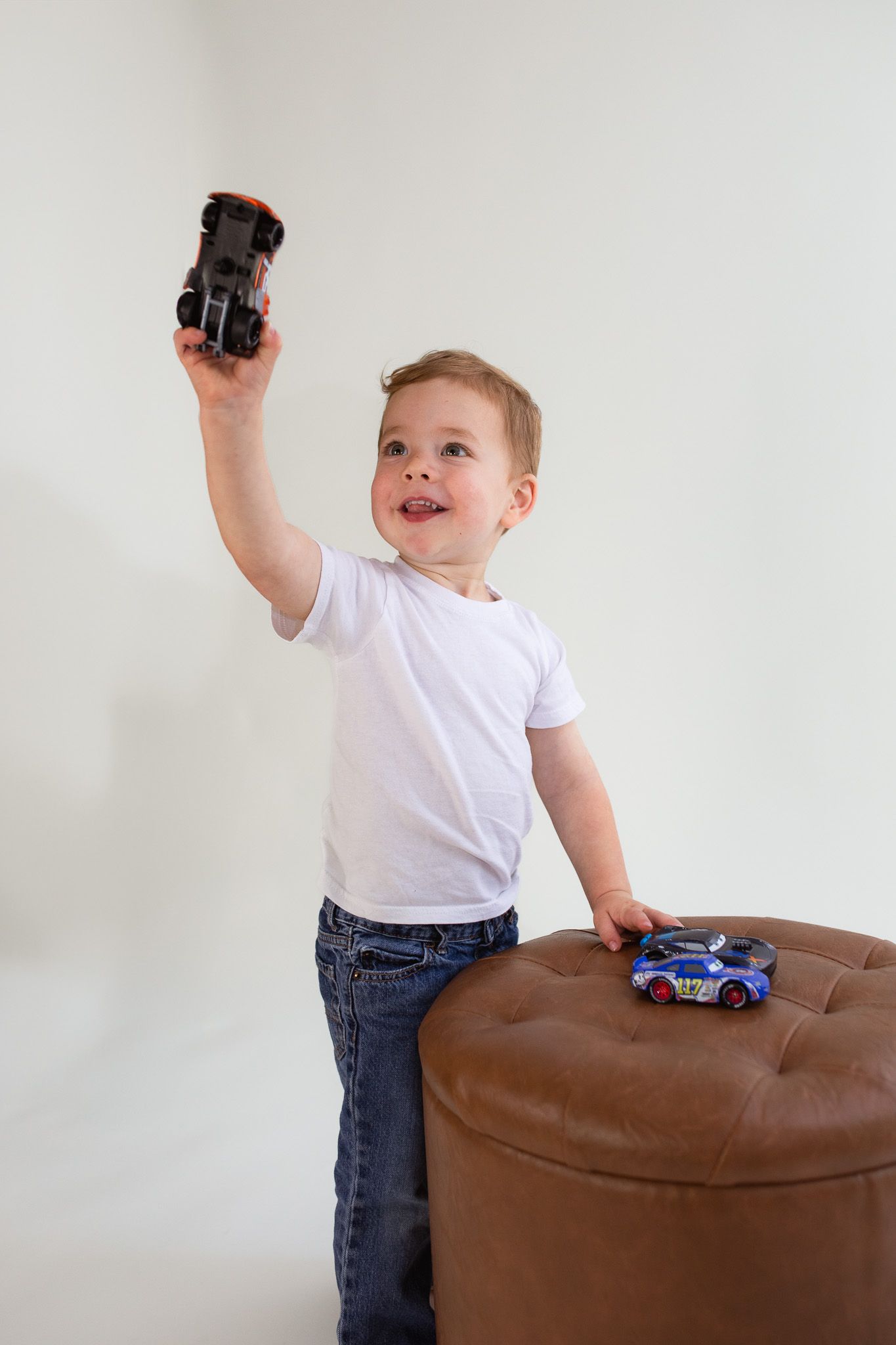 A young child in white shirt holding up a toy vehicle next to a brown ottoman with another toy car on top.