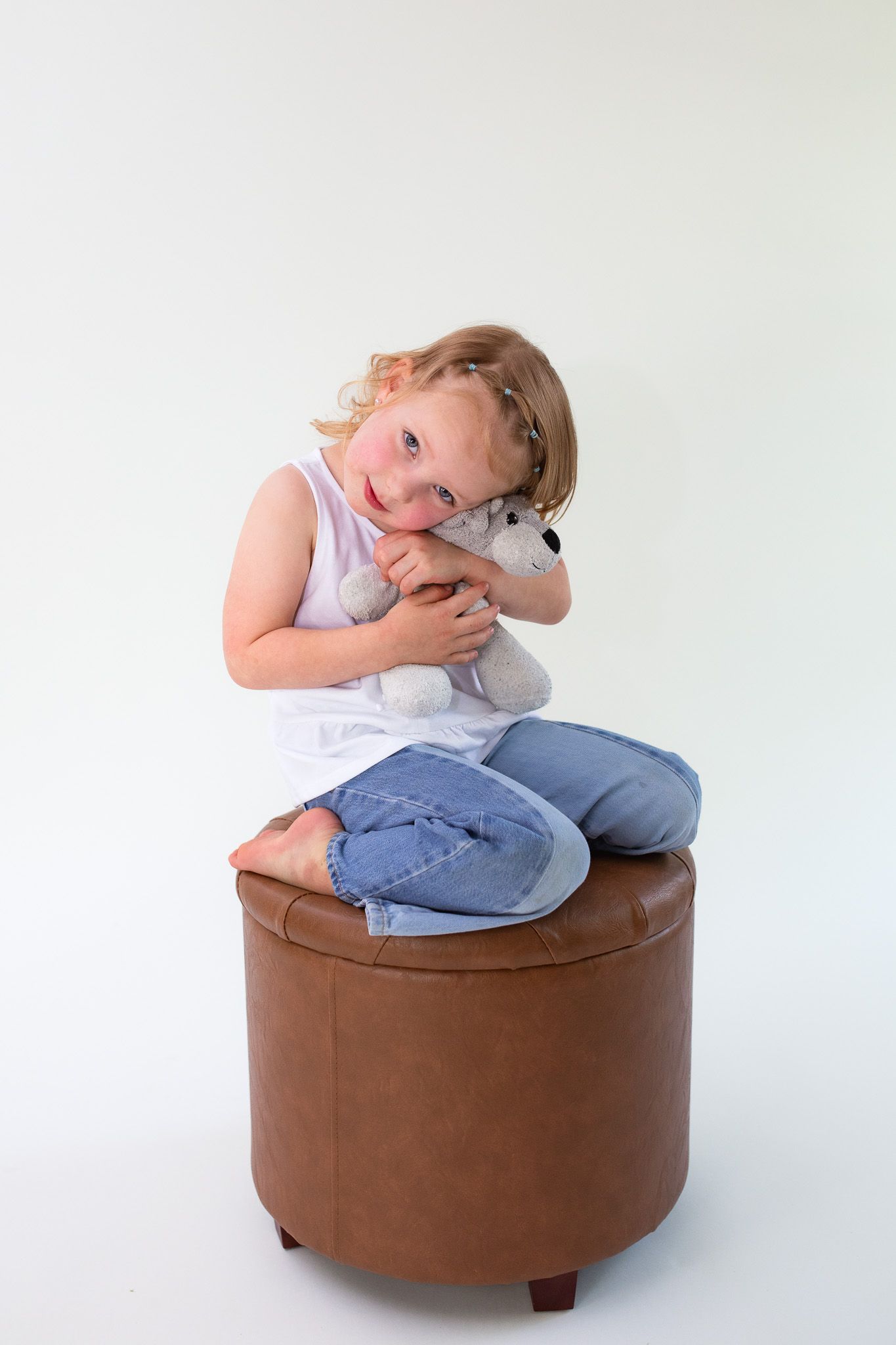 A young child wearing jeans and a white tank top hugs a stuffed animal while sitting on a round brown ottoman.
