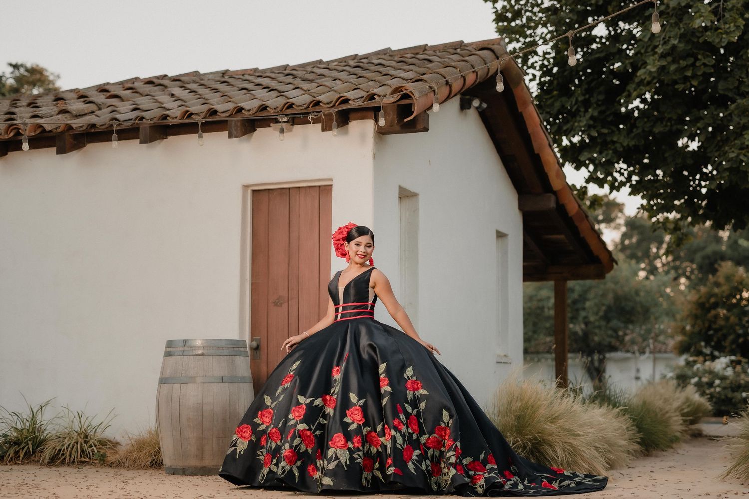 A quinceanera in an elegant black and red floral ball gown stands beside a rustic white adobe building with a barrel.