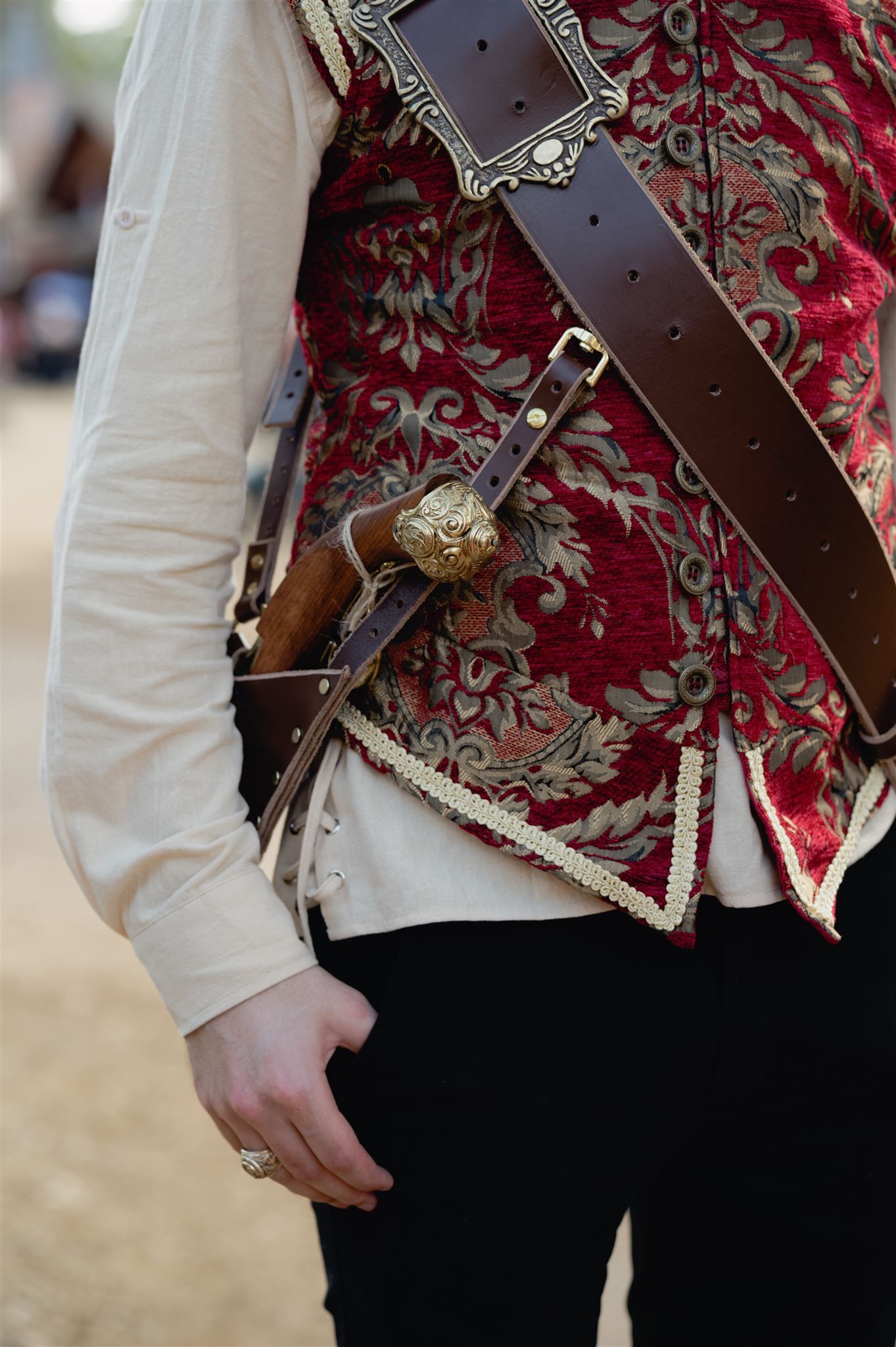 Close-up of a brown leather belt and ornate red brocade vest with floral patterns worn over a white shirt.
