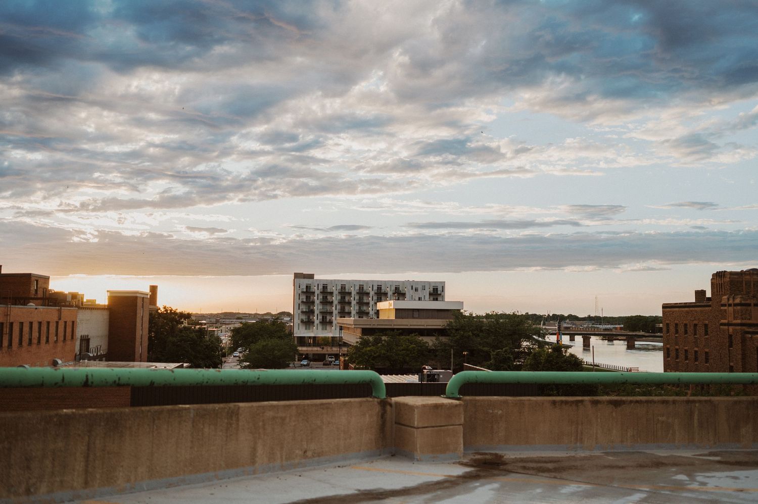 A cityscape view at sunset with turquoise pipes on a concrete rooftop and dramatic cloudy skies overhead.