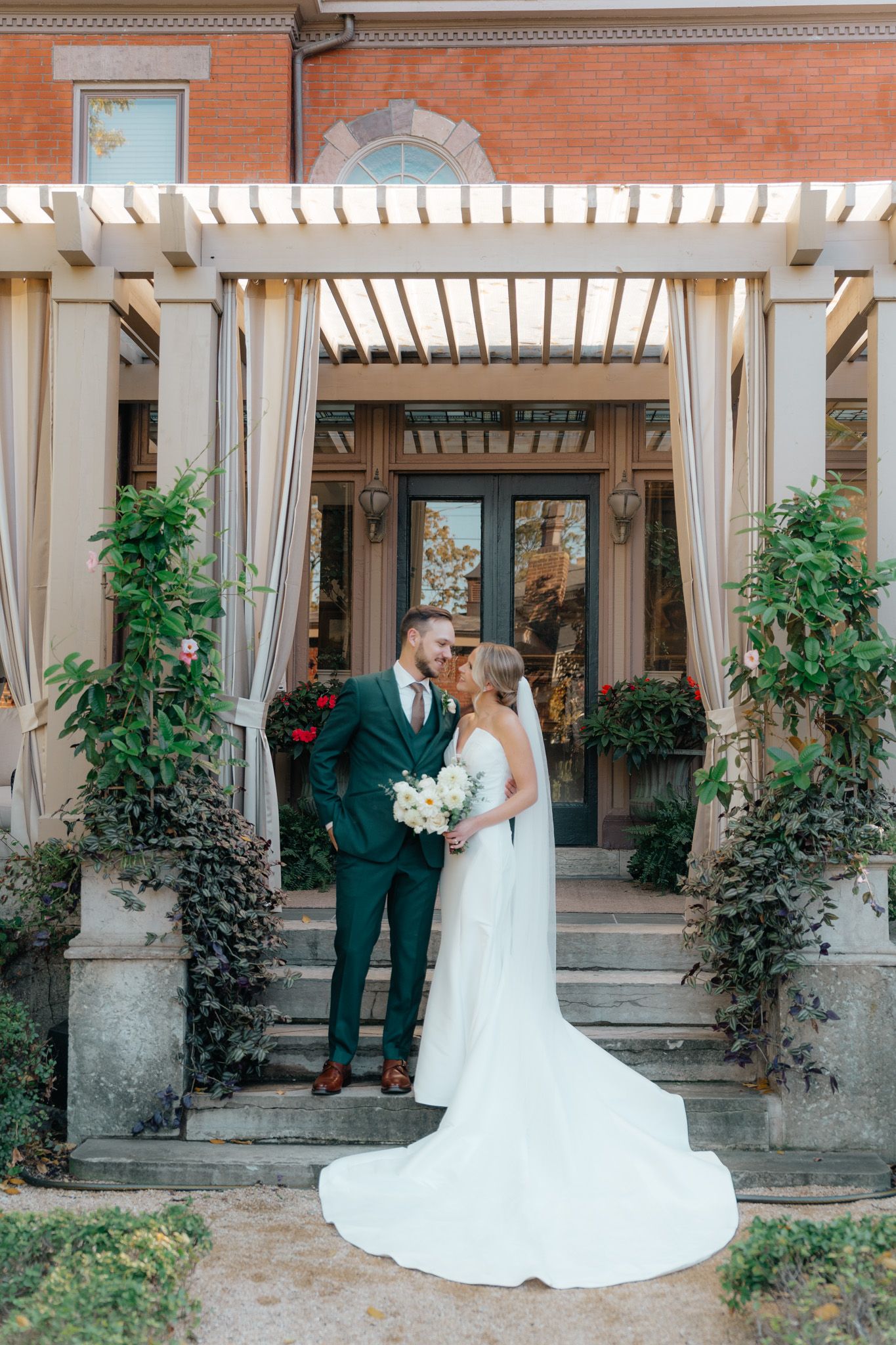 Wedding couple in elegant attire standing under pergola entrance of brick building.
