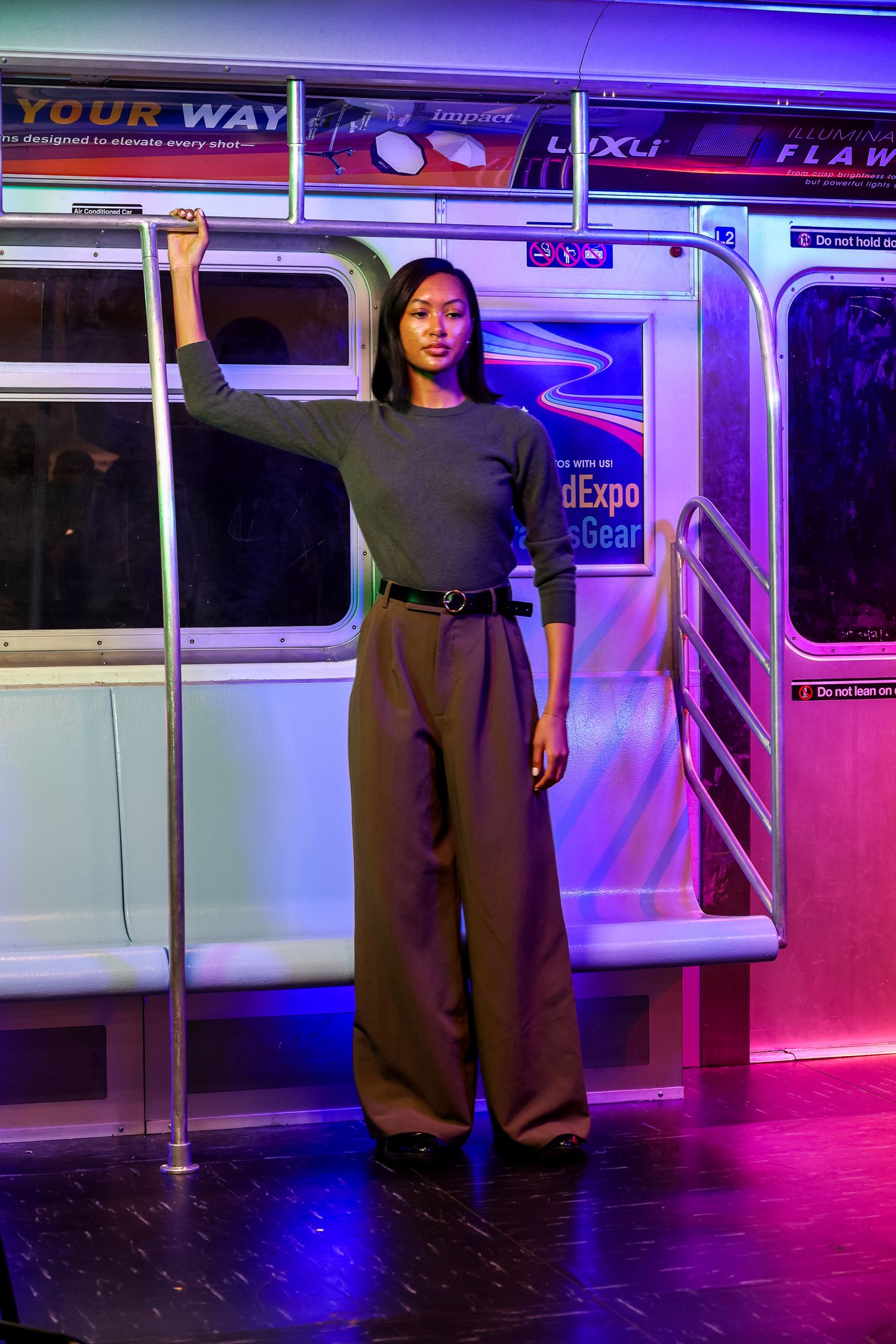 A person poses confidently on a subway train wearing a black top and wide-leg pants with neon lighting in the background.