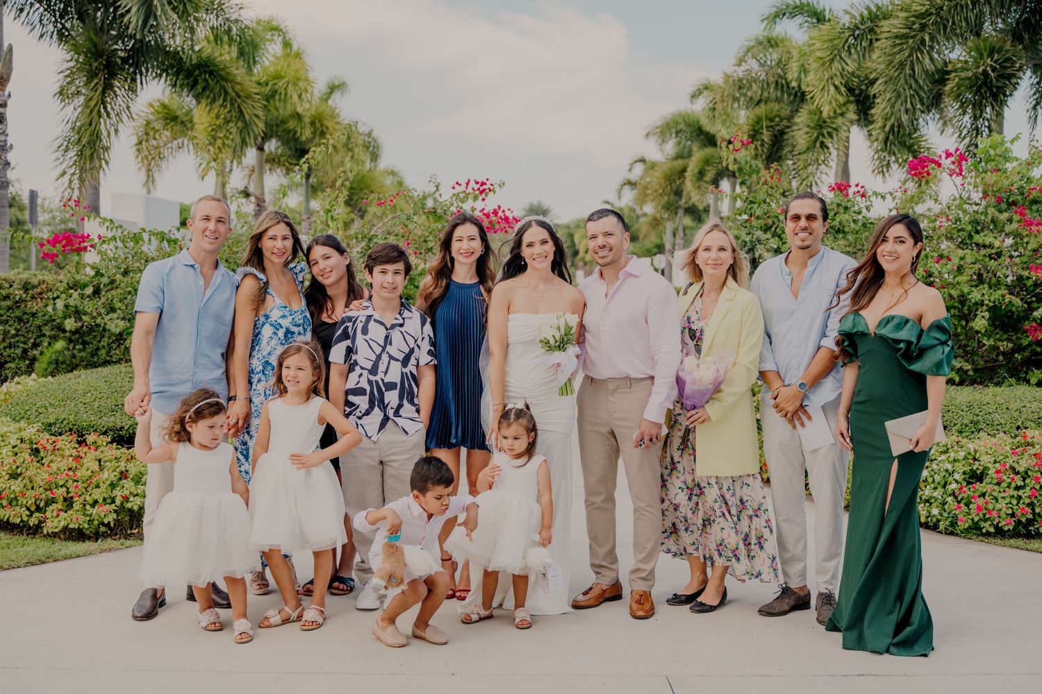 Wedding party members in light colored attire pose together in a tropical garden setting with palm trees and flowers.