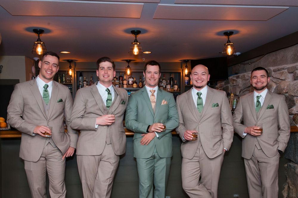 A wedding party poses together wearing matching tan and sage green suits in a formal indoor setting.