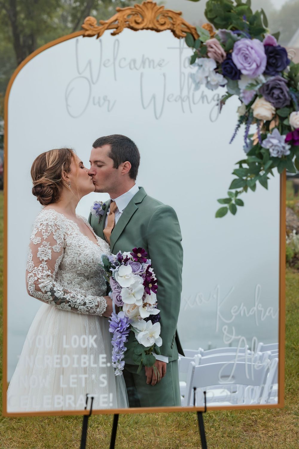 Wedding portrait in front of an ornate gold mirror with purple floral decor and a welcome sign display.