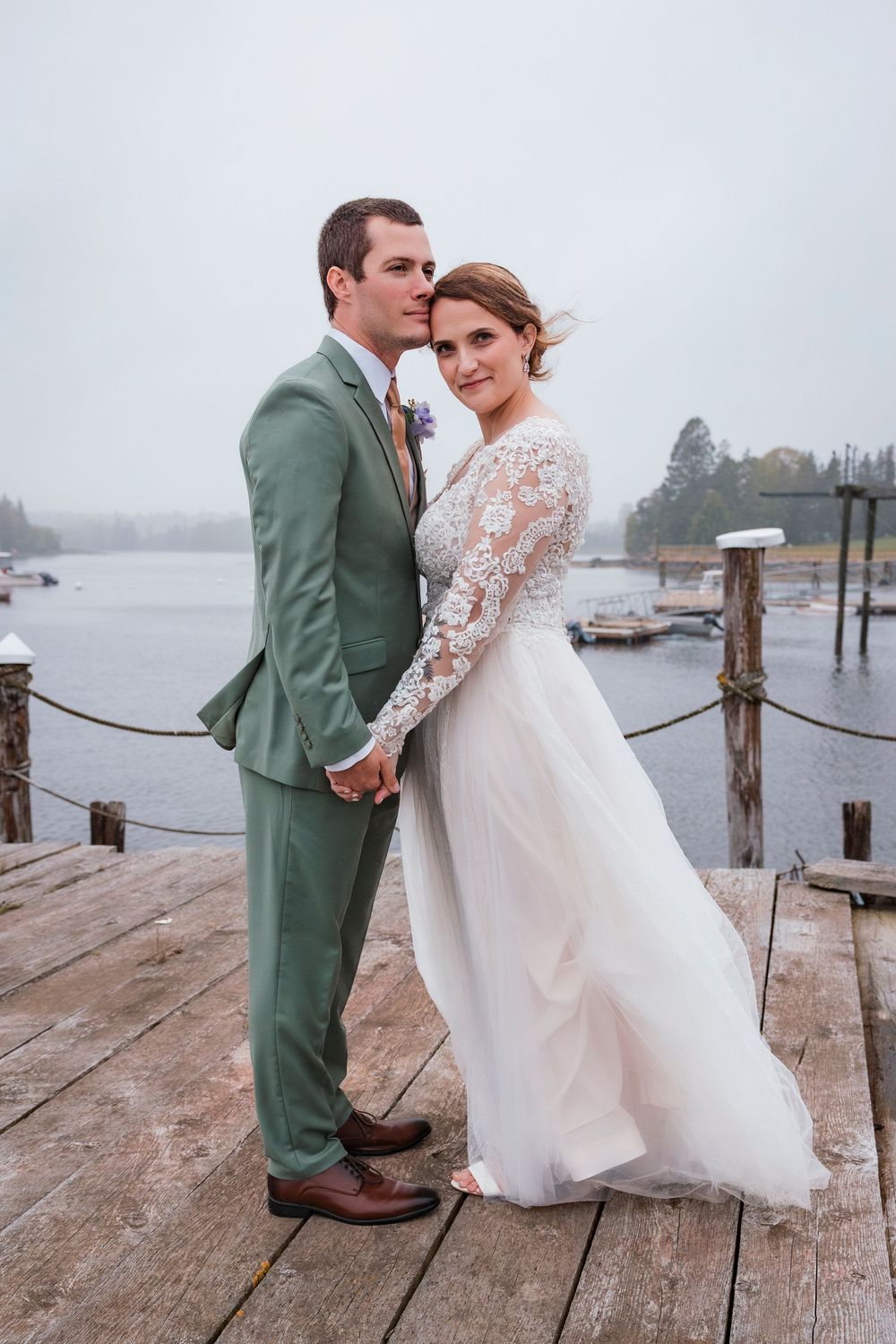 A couple in wedding attire stands on a wooden dock overlooking a misty waterfront.