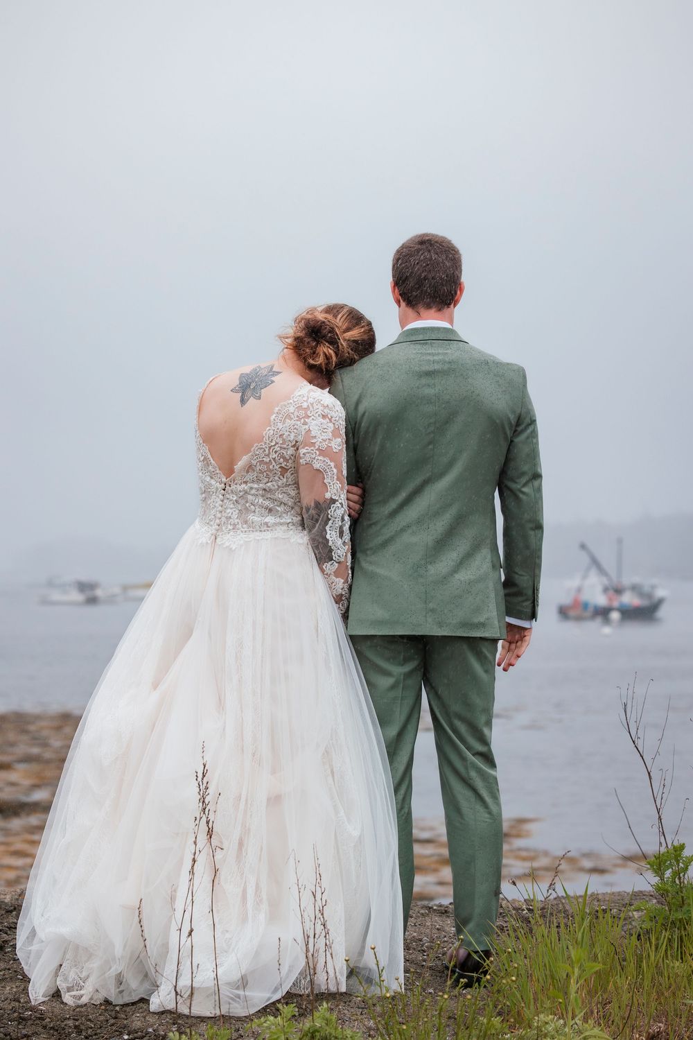 A couple in wedding attire stands at the edge of a misty harbor with boats visible in the background.