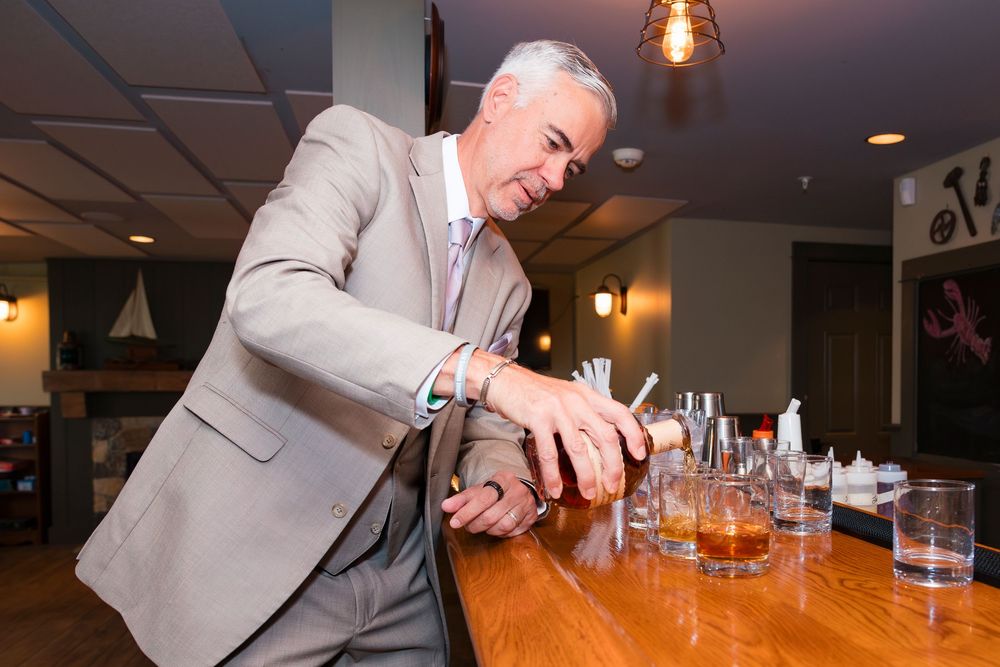 Someone in a beige suit pours a cocktail at a wooden bar counter with multiple glasses lined up.