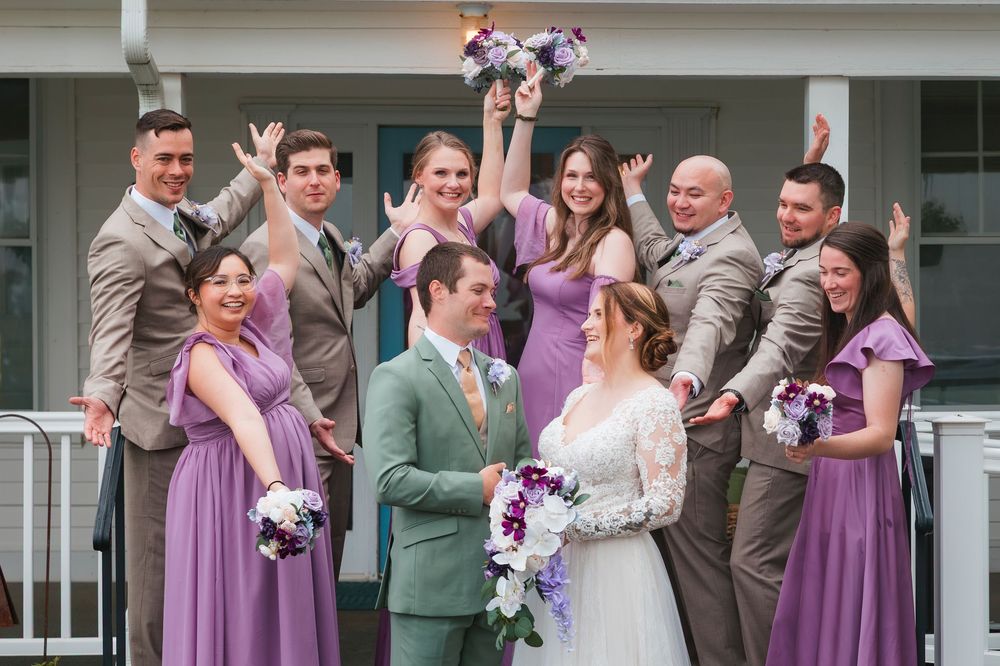 Wedding party wearing purple bridesmaid dresses and gray suits laughs together outdoors during photo session.