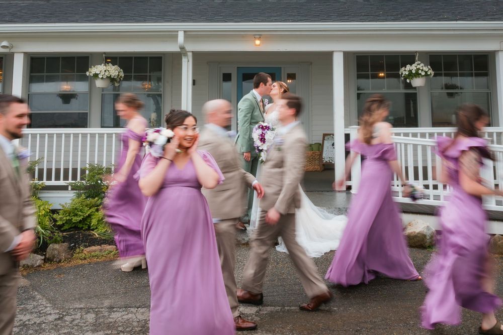 Motion-blurred wedding party in purple bridesmaid dresses and tan suits walks outside a house with white porch columns.