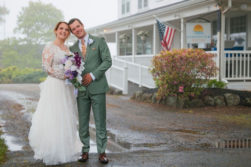 A couple in wedding attire stands together on a rainy day outside a building with an American flag.