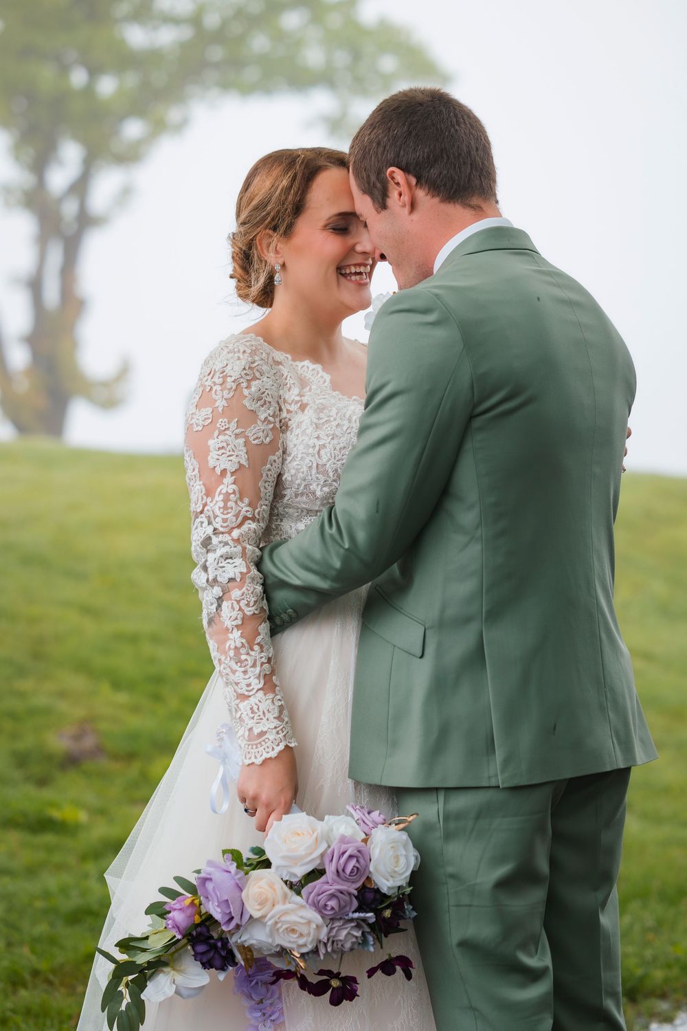 A romantic wedding moment captured outdoors with a beautiful lace dress and sage green suit against a misty natural backdrop.