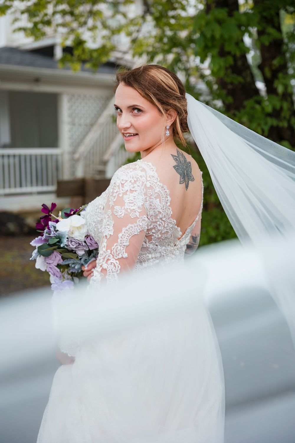 A flowing white wedding veil billows in the breeze as a bride poses with her purple and white floral bouquet.