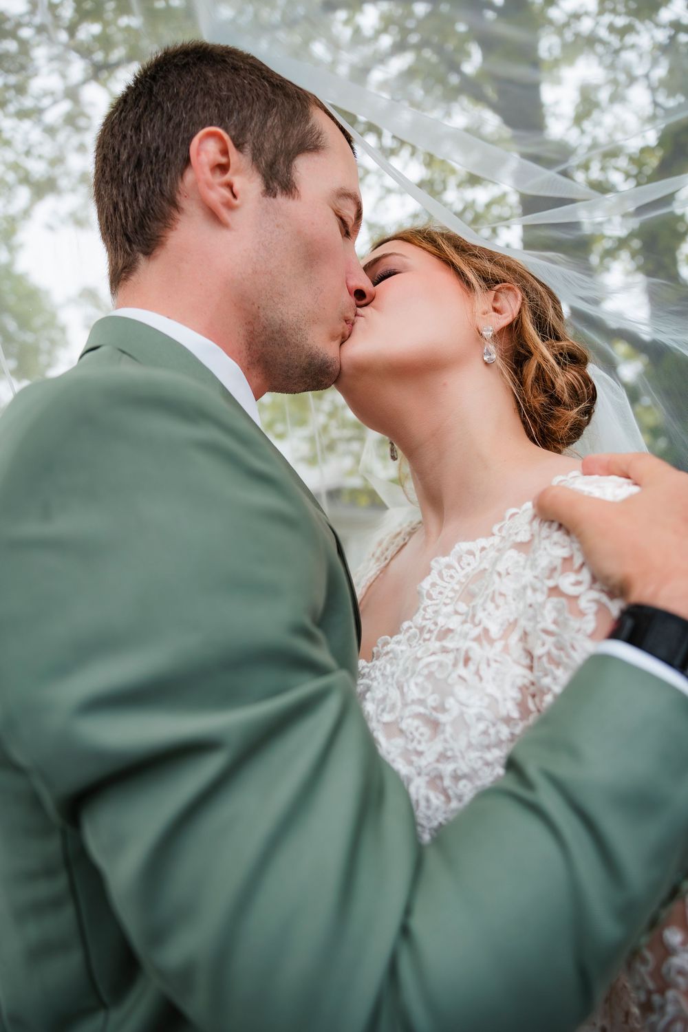 A romantic wedding couple sharing a kiss outdoors with soft lighting and a sage green suit.
