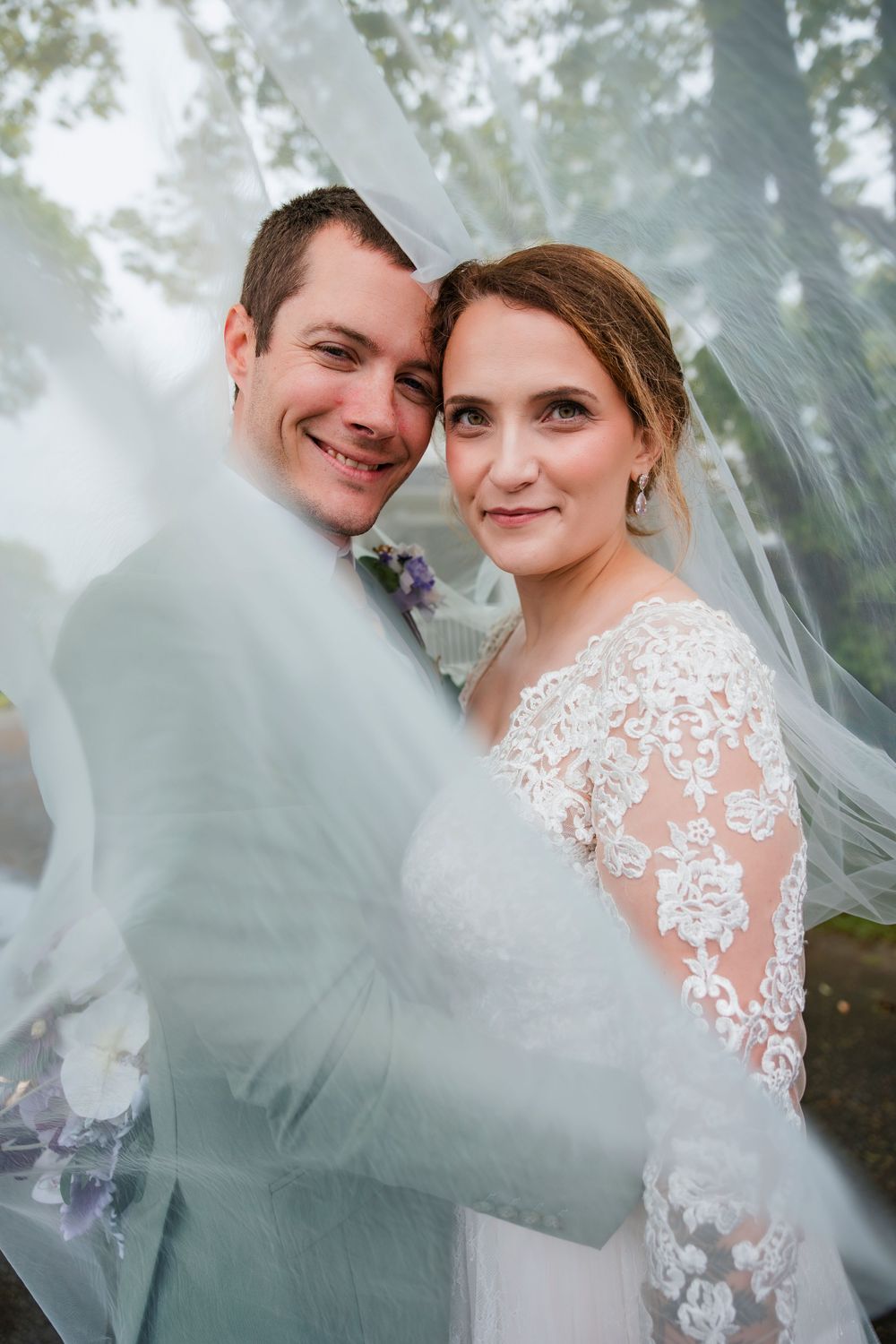 A romantic wedding portrait through a sheer veil showing a couple embracing outdoors.
