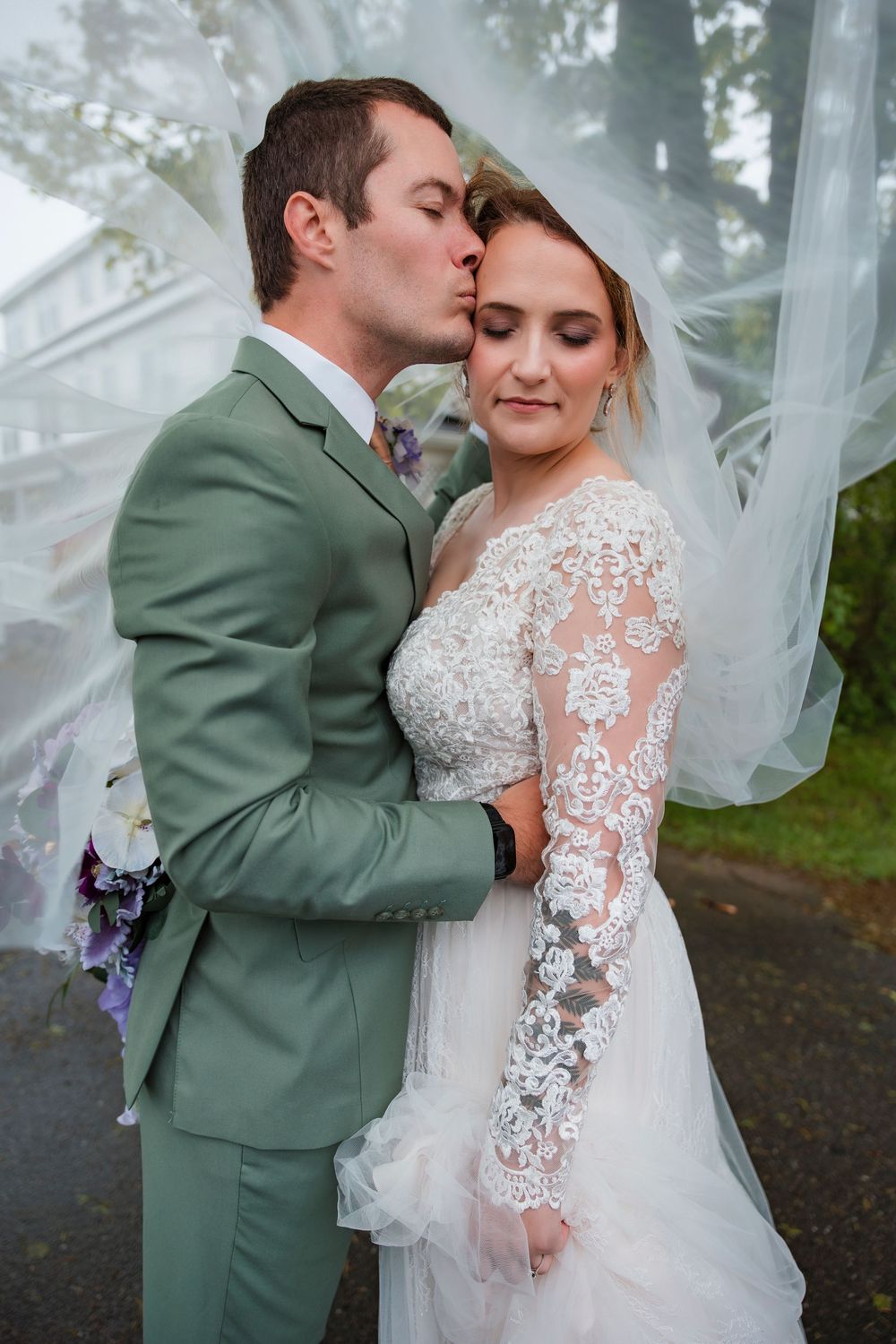 A tender wedding moment captured with the groom in sage green suit kissing the bride in a lace gown.