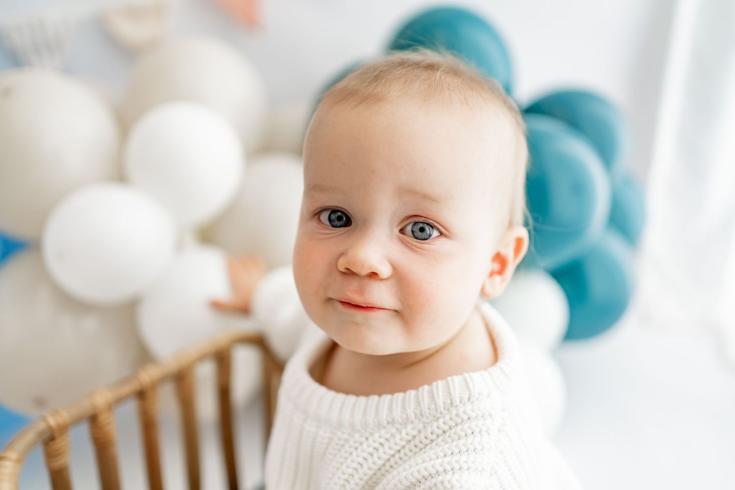 A baby surrounded by blue and white balloons sits on a wooden chair during a special occasion.