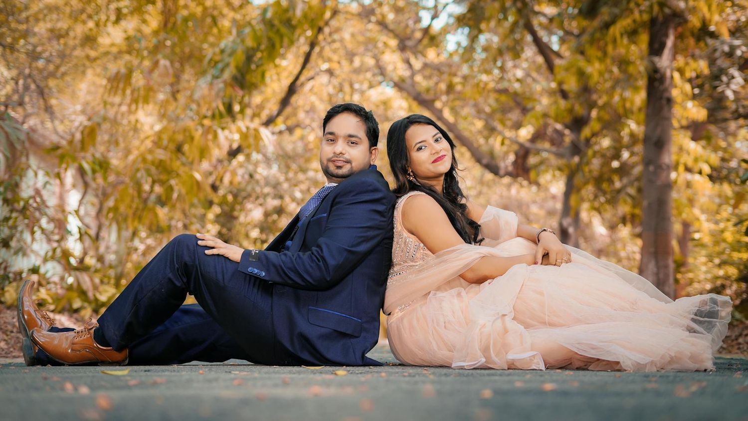 A couple sits back-to-back on a stone surface surrounded by golden autumn trees in an outdoor setting.