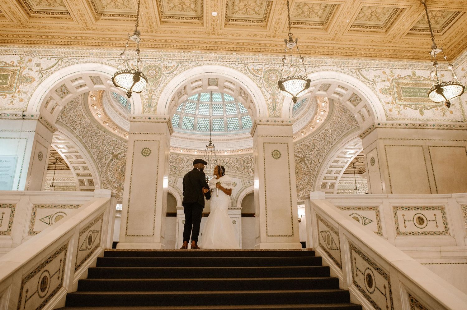Ornate white marble staircase with gilded coffered ceiling and elegant chandeliers in a grand Beaux-Arts style interior.