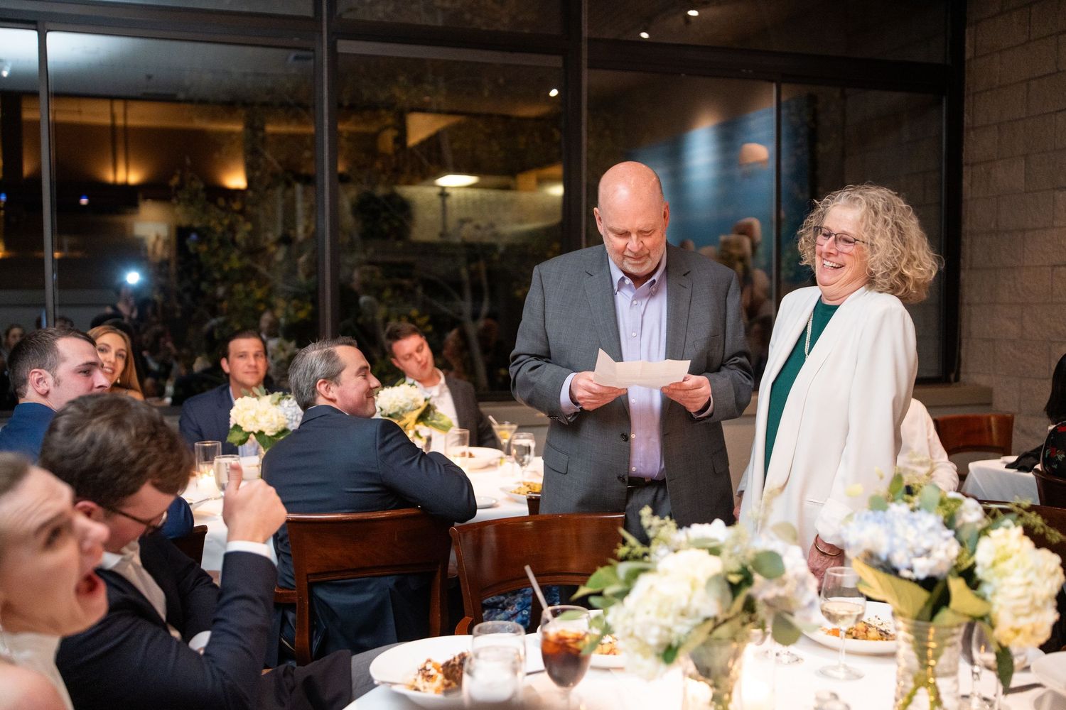 Elegantly dressed guests at a dinner party stand around a table with white floral centerpieces in an upscale restaurant setting.