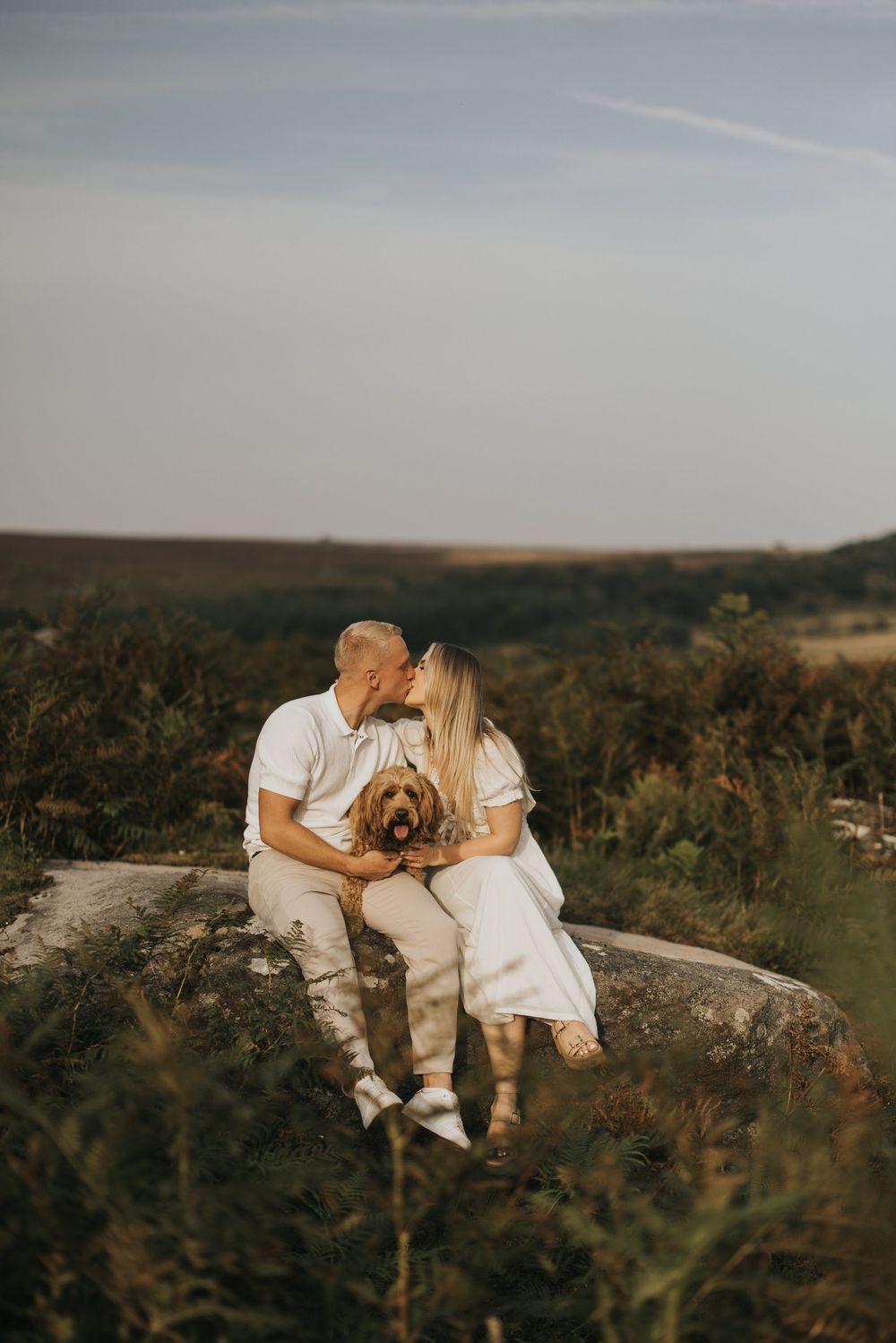 A couple in white outfits embraces on a hilltop at sunset among wild desert shrubs during an engagement photoshoot.