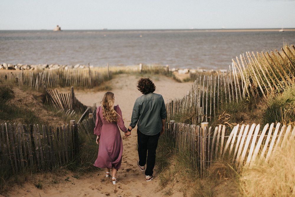 Two people walk along a beach path with wooden fences and tall grass against a cloudy coastal backdrop.